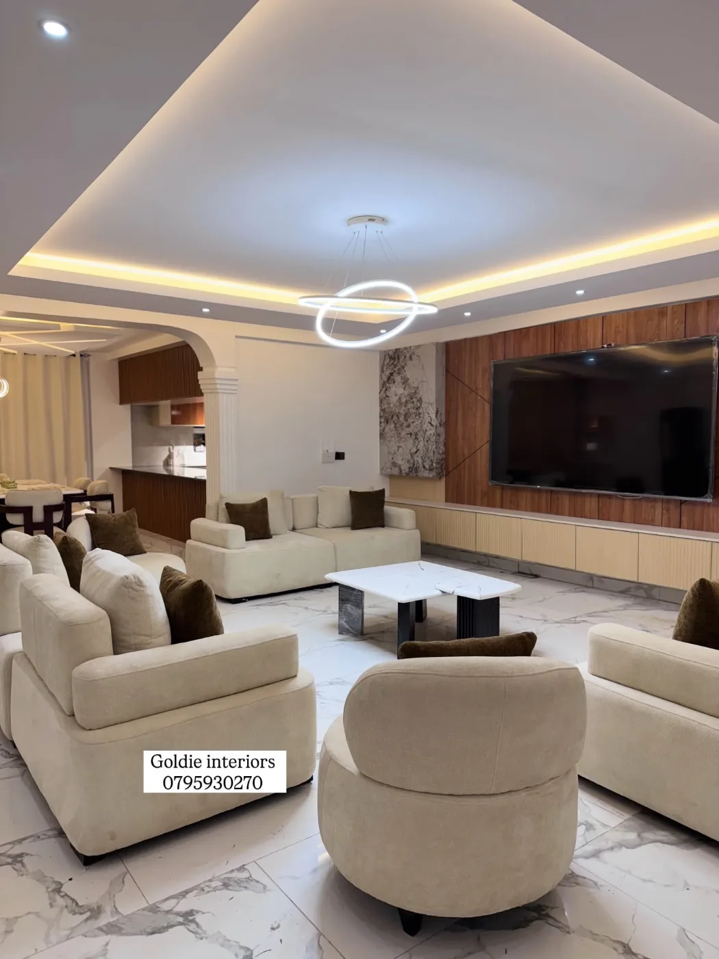 Living room with cream sofas and a marble-top coffee table facing a wall-mounted TV on a timber-slat feature wall, under a gypsum ceiling with warm edge lighting and a ring chandelier