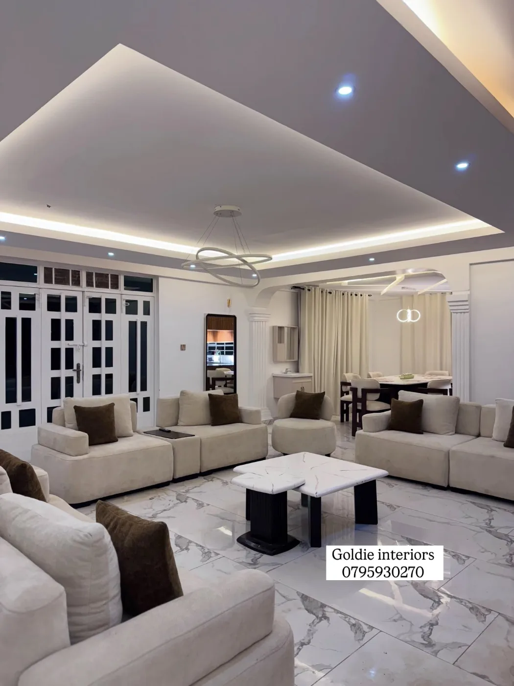 Wide view of the living room with cream sofas and brown cushions, white double doors, an arch to the dining area and a stepped ceiling with concealed lighting