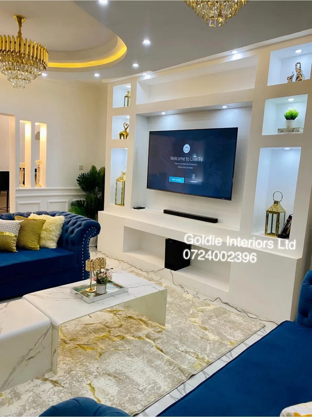 Built-in white TV unit with lit alcoves, a wall-mounted TV and sound bar, next to the blue sofa