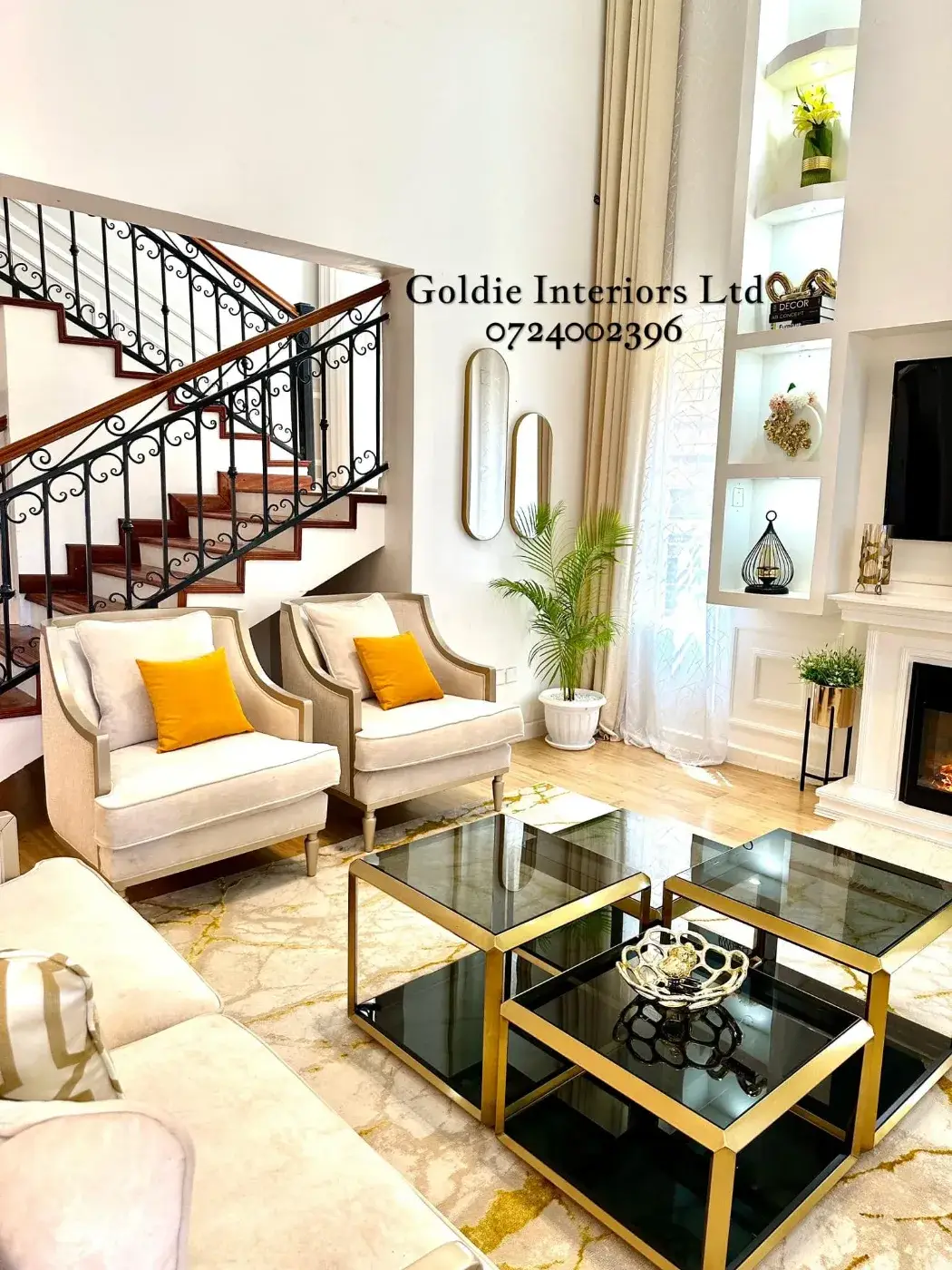 Double-height living room with cream armchairs and mustard cushions, nested gold-framed glass coffee tables, a wrought-iron staircase, oval gold mirrors and lit white wall niches beside the fireplace