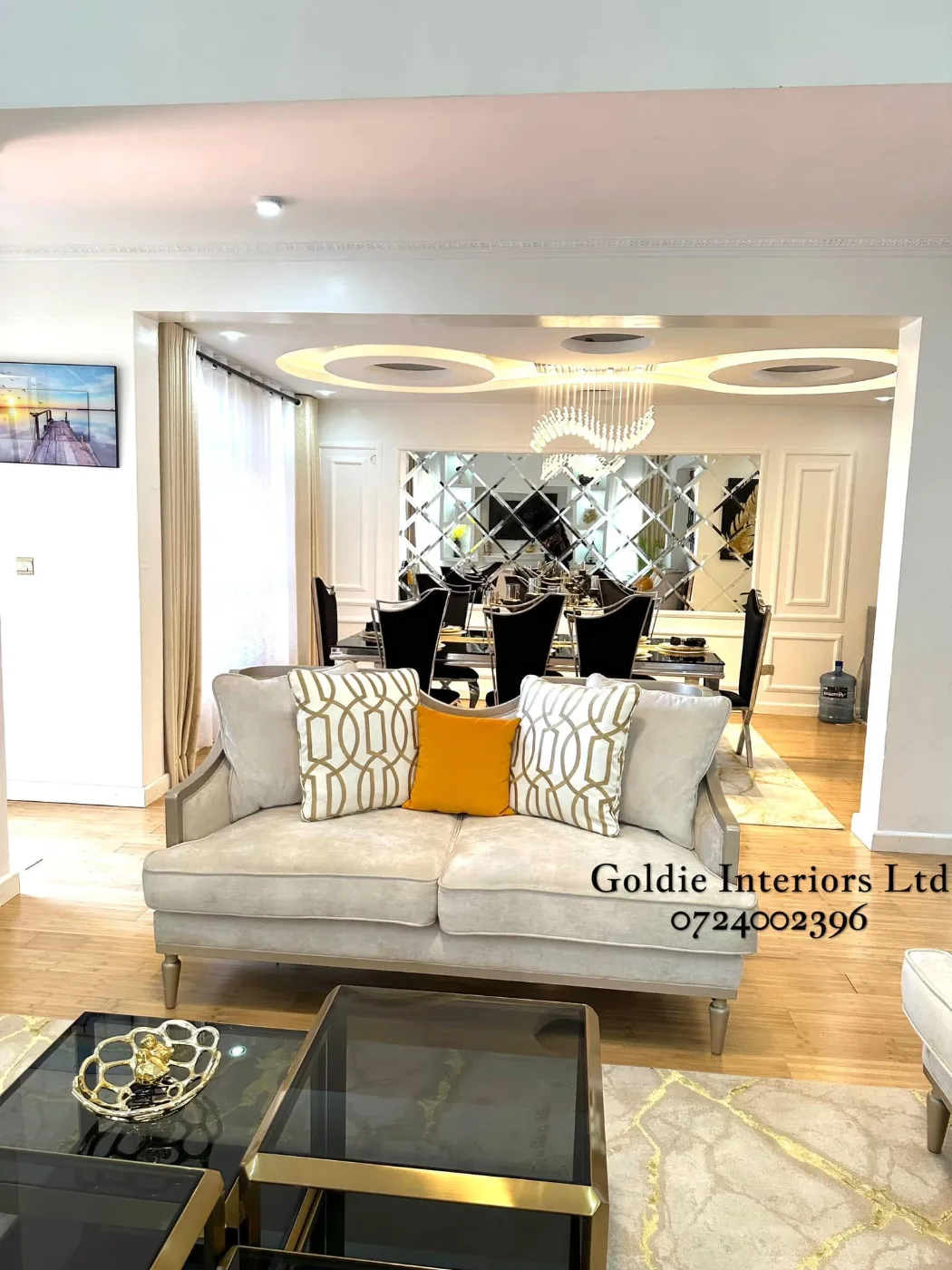 Cream sofa with gold-patterned and mustard cushions in front of the dining area, with a looping gypsum ceiling, a crystal chandelier and a bevelled mirror wall