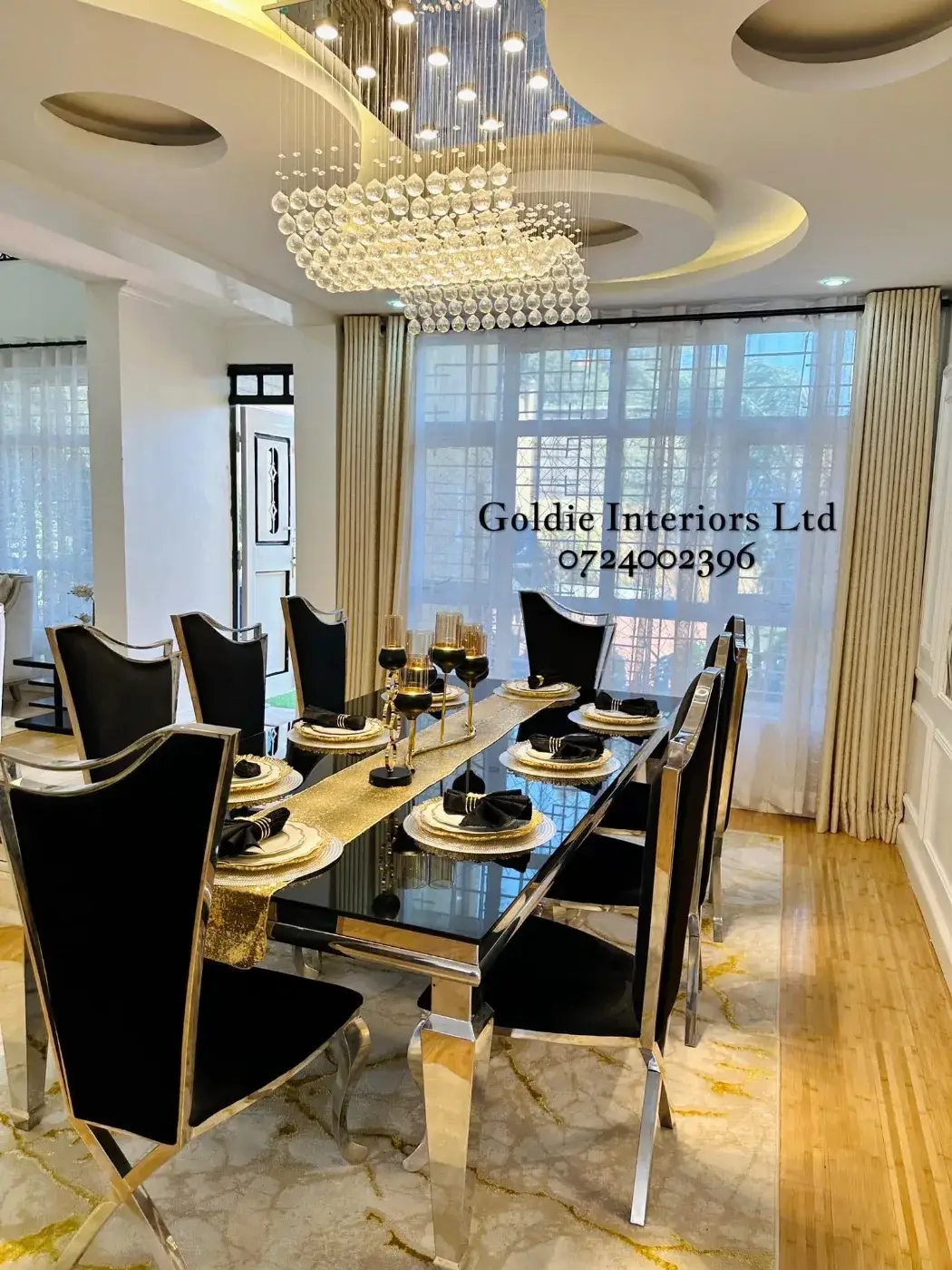 Black-and-gold dining table set for eight with high-backed black chairs, a gold runner and candle holders, under a gypsum ceiling with a cascading crystal chandelier
