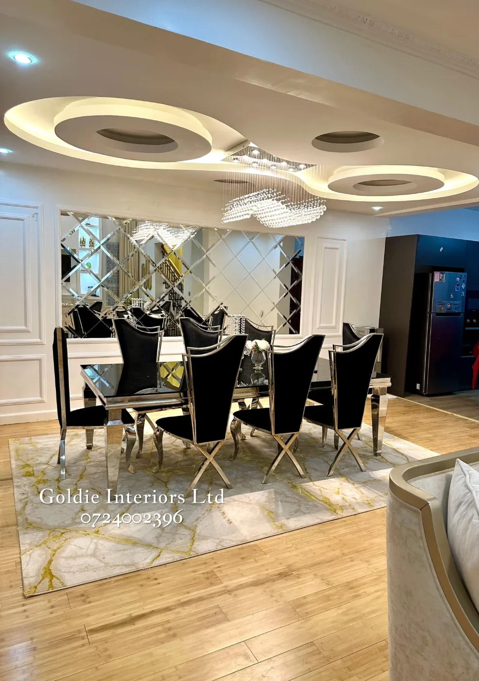 Dining room with a bevelled-edge mirror panel between white wainscoting, black chairs with metal frames on a cream-and-gold rug and a curved lit gypsum ceiling