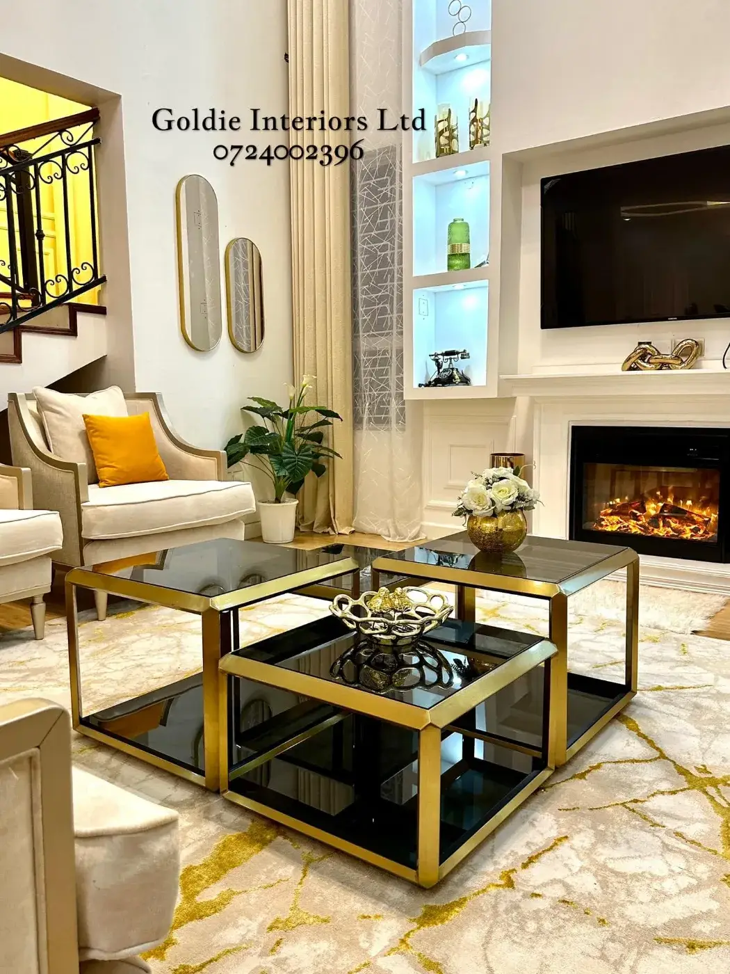 Gold-framed glass coffee tables in front of the electric fireplace, a wall-mounted TV and lit white niches, with oval mirrors and the staircase to the left