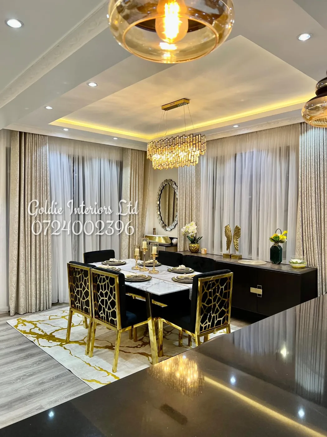 Dining table with gold-framed black chairs beside a black sideboard and counter, under pendant lights and a lit ceiling