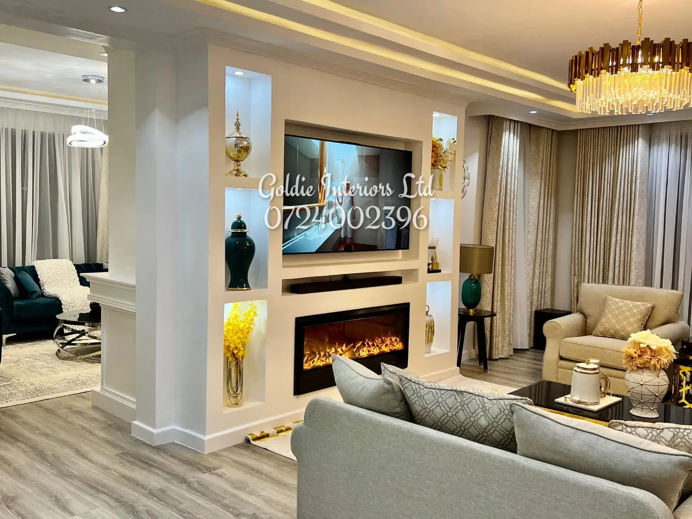 Living room with a lit TV wall, electric fireplace, grey sofa and chandelier beside a white column