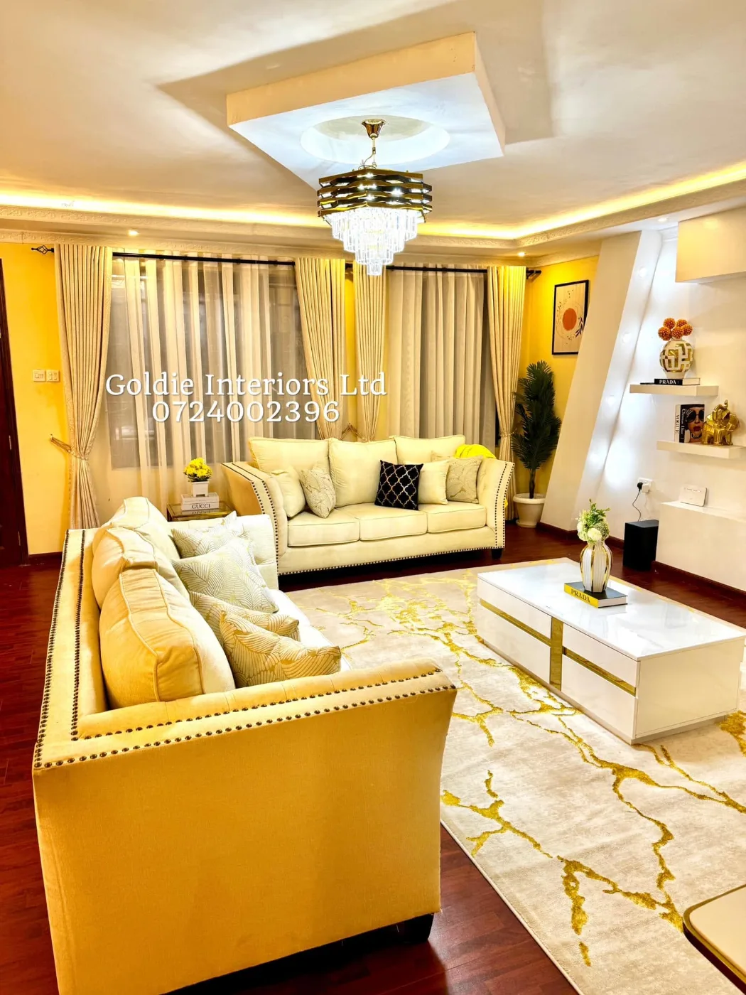 Living room with cream studded sofas, a white coffee table with gold trim on a cream-and-gold rug, a crystal chandelier and cream curtains with sheers