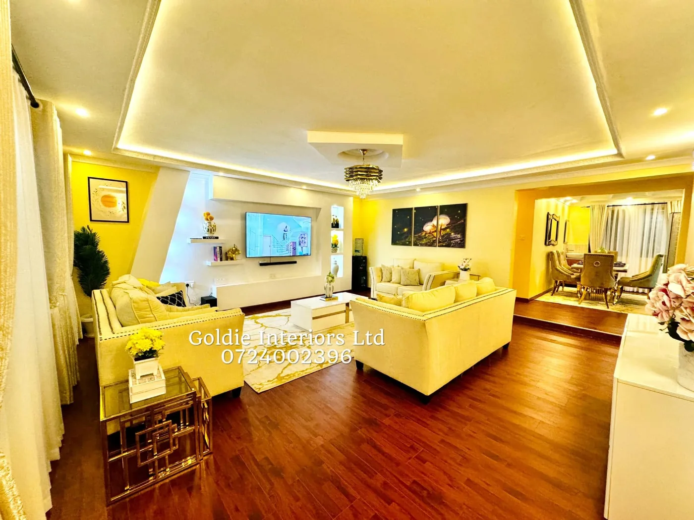 Wide view of the open living room with cream sofas, gold-framed side tables, a white TV unit and a lit gypsum ceiling, with the dining area at the far end