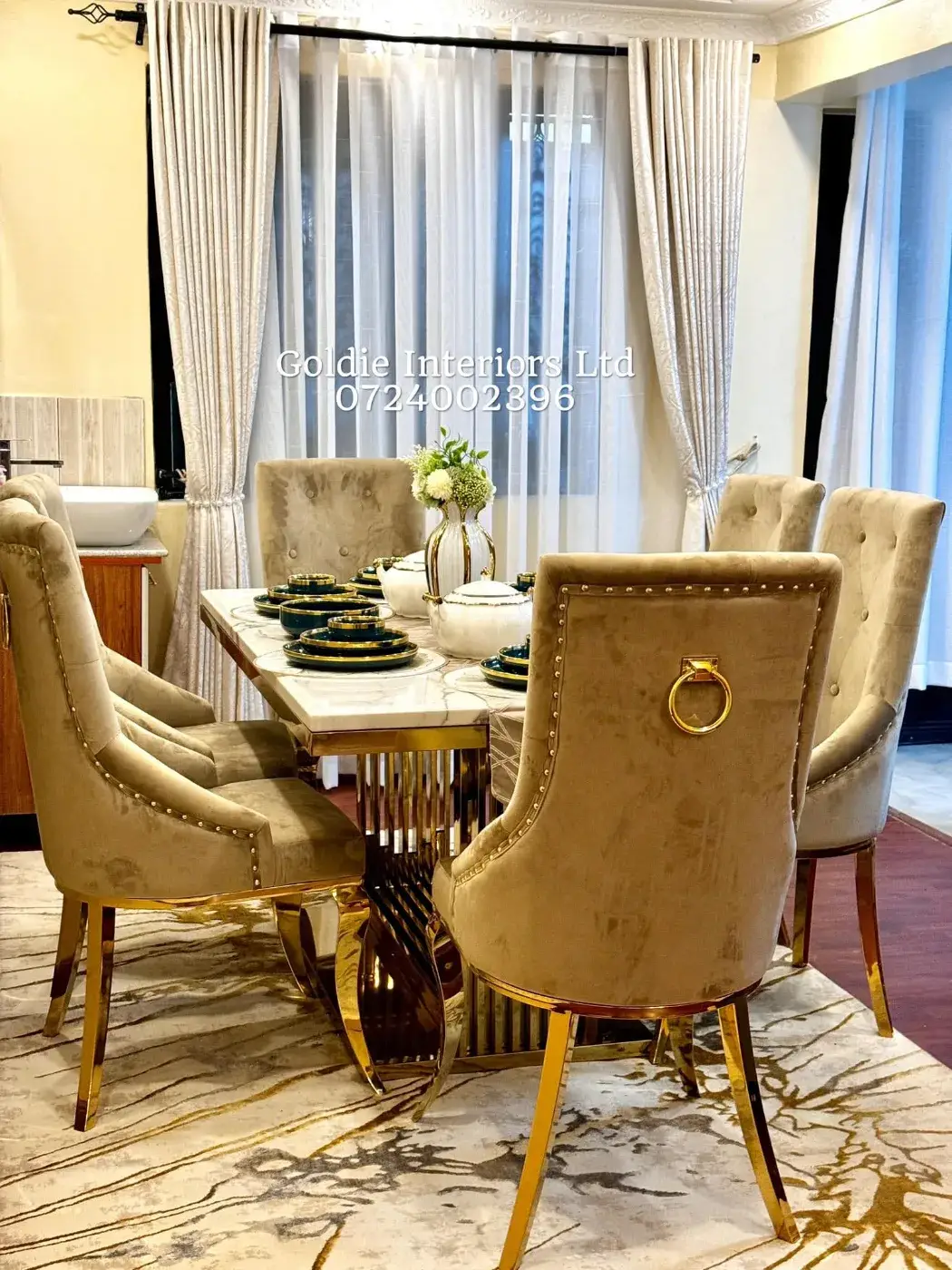 Dining table set with teal and gold crockery, surrounded by tufted taupe velvet chairs with gold legs and ring handles, in front of sheer and patterned curtains