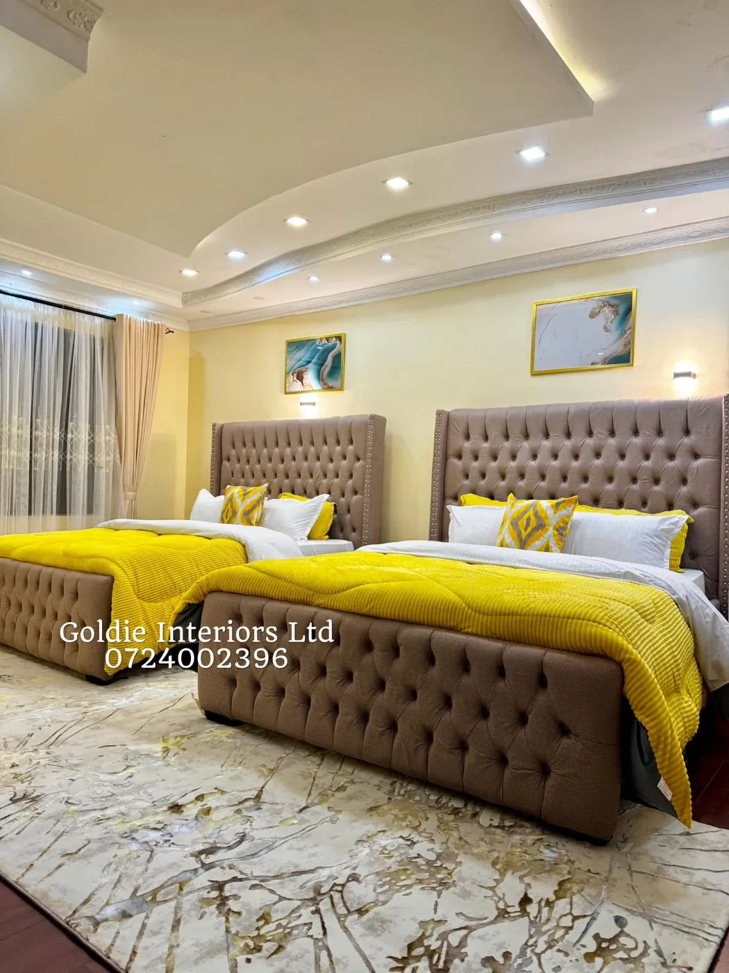 Bedroom with two tufted taupe beds, yellow throws, white pillows, patterned cushions and a cream-and-gold rug beneath a curved ceiling