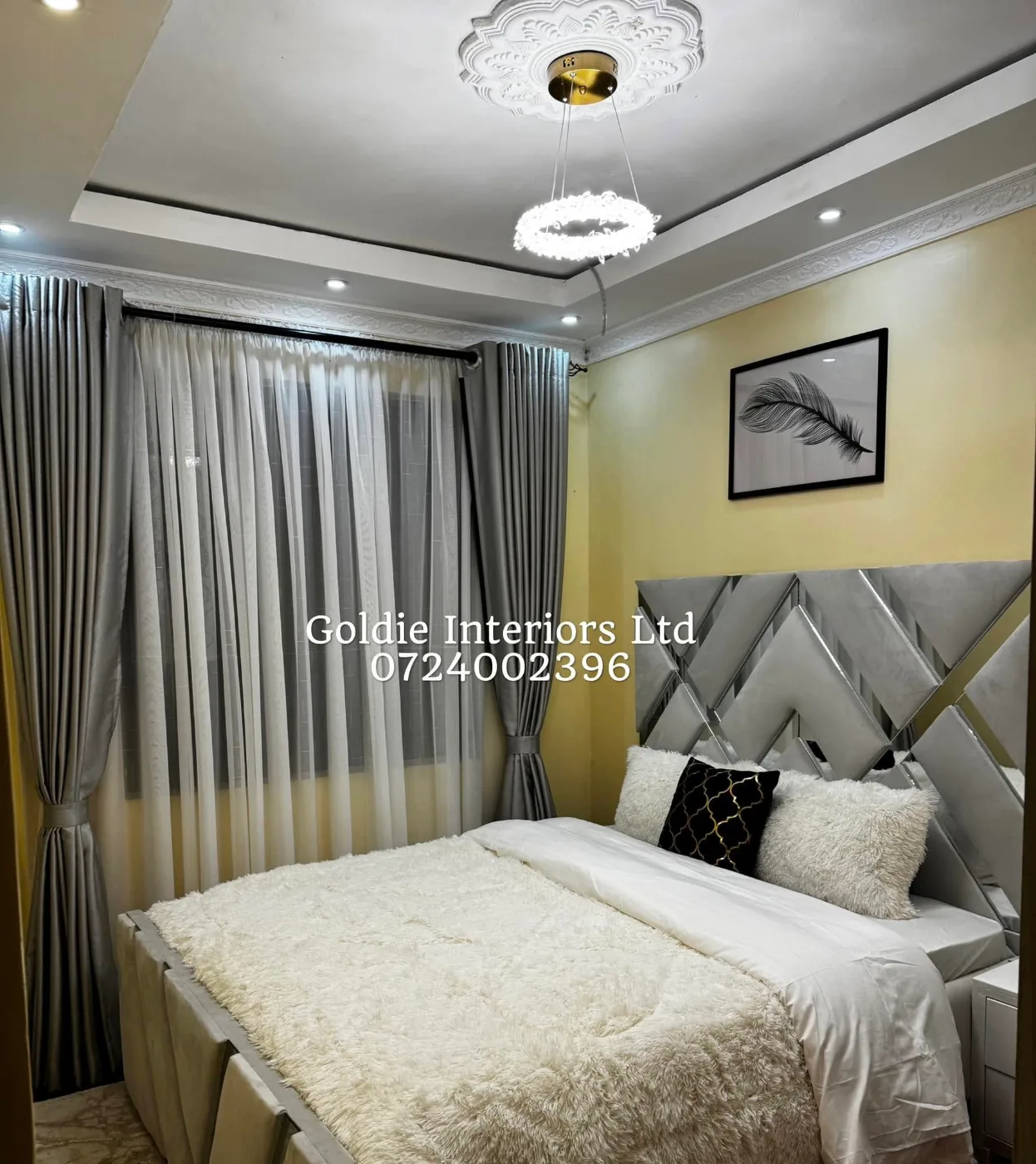 Bedroom with a grey panelled headboard with mirrored edging, white fur throw and pillows, grey curtains with white sheers and a ring chandelier